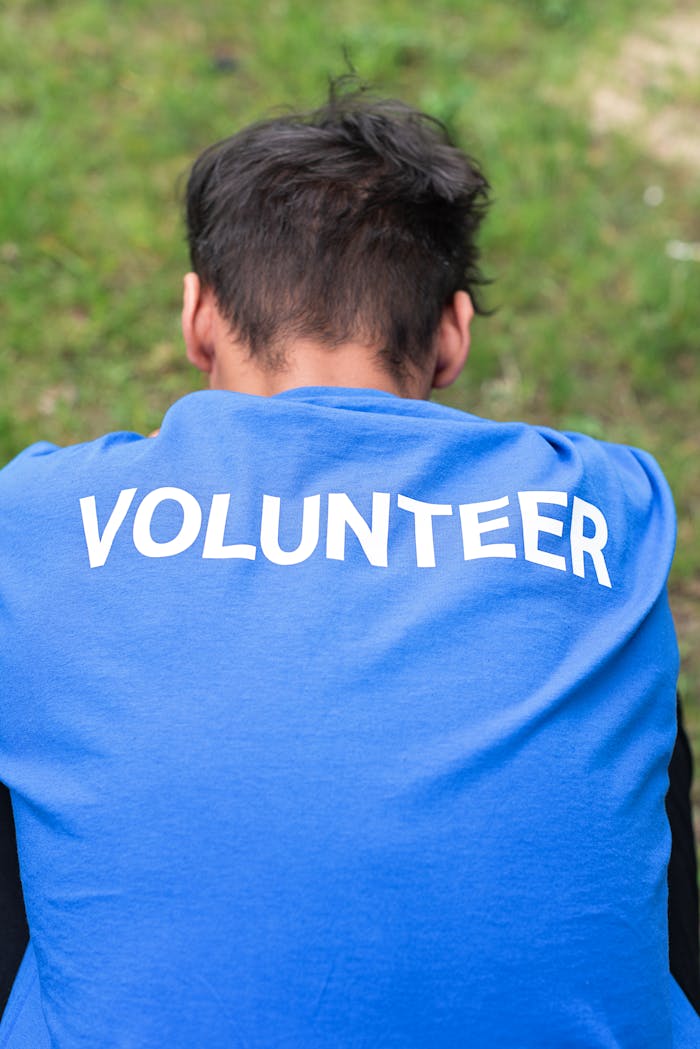 Man wearing a blue volunteer shirt sits on grass, photographed from behind.